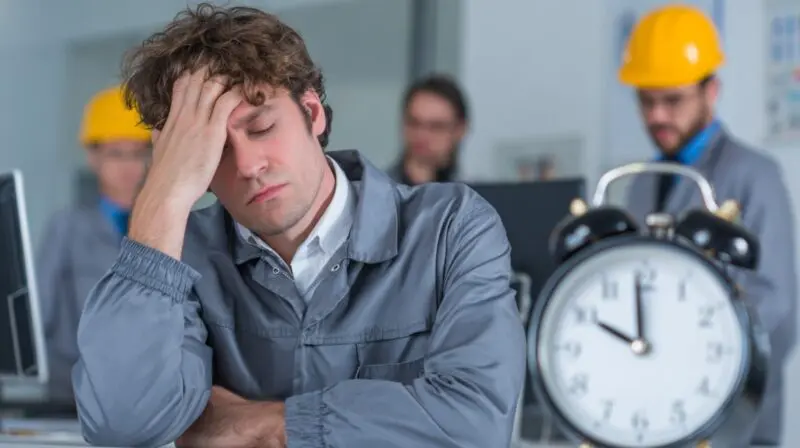 Tired worker sitting at a desk with coworkers and a clock in the background